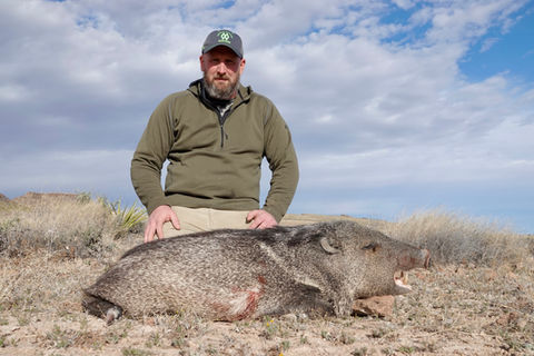 A hunter in a green sweatshirt kneels beside the javelina he has bagged