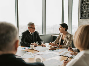 Business meeting with two professionals discussing documents at a table. Papers with charts, cup, and pens. Large windows in the background.