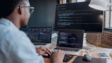 Person coding at a desk with two laptops and a large monitor displaying code. Brick wall and window in the background; focused atmosphere.
