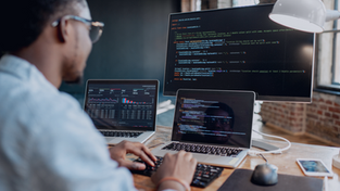 Person coding at a desk with two laptops and a large monitor displaying code. Brick wall and window in the background; focused atmosphere.