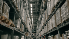 Tall warehouse shelves filled with pallets wrapped in plastic. Bright ceiling lights and signs are visible. It's a tidy, industrial setting.