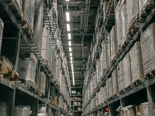 Tall warehouse shelves filled with pallets wrapped in plastic. Bright ceiling lights and signs are visible. It's a tidy, industrial setting.
