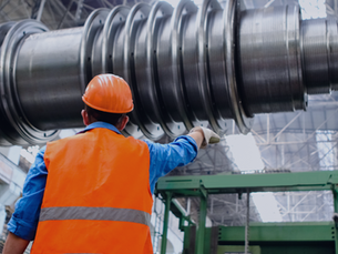Worker in orange vest and helmet examines large metal machinery in an industrial facility. High ceiling with metal framework above.