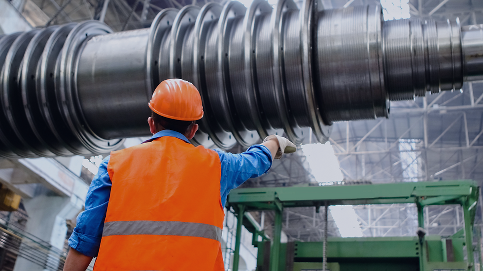 Worker in orange vest and helmet examines large metal machinery in an industrial facility. High ceiling with metal framework above.