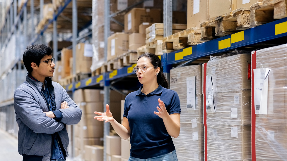 Two people in a warehouse discuss logistics. One gestures while speaking, surrounded by shelves of cardboard boxes. Blue shelves add contrast.