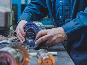 Hands inspecting a large metal bearing on a workbench, surrounded by gears and tools. Person wears a blue coverall, indicating a workshop setting.