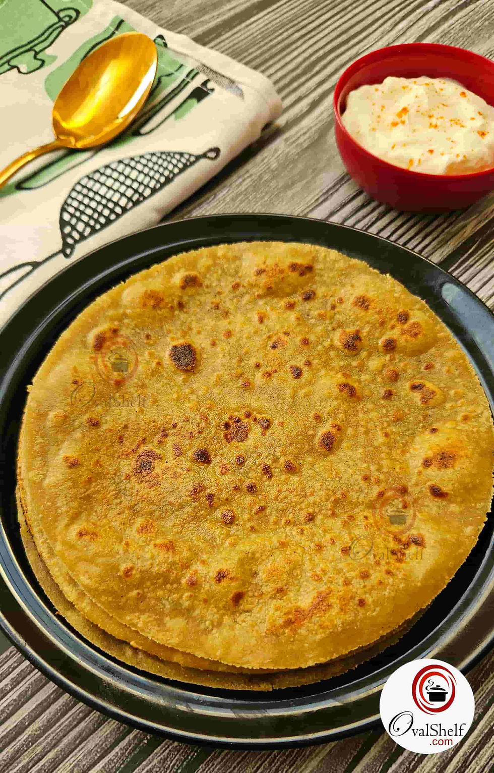 Stacked Quinoa flatbreads / Rotis on a black plate with a red bowl of yogurt, a gold spoon, and a patterned napkin on a wood table.