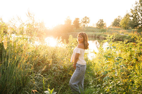 A high school aged woman with light brown hair wearing blue jeans and an off the shoulder white top outside near a pond with reeds and trees in the background