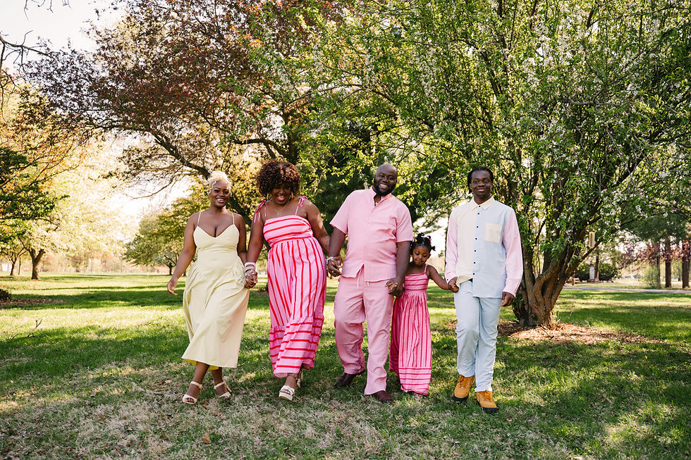 A joyful family of five walks hand-in-hand through a park. They wear colorful outfits—pink, yellow, and white—amidst lush green trees.