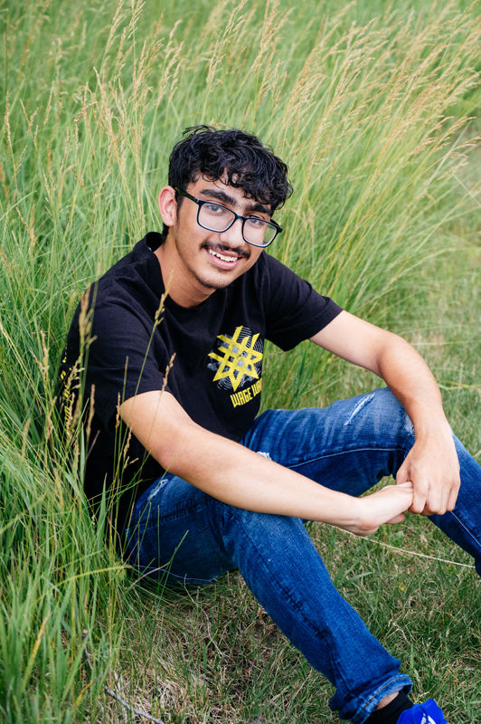 Young man sitting in tall grass, smiling, wearing glasses, a black graphic tee, and jeans. Relaxed setting with vibrant green background.