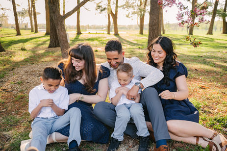 A family of five sits on grass in a sunlit park, laughing together. Tall trees and pink blossoms surround them, creating a warm atmosphere.