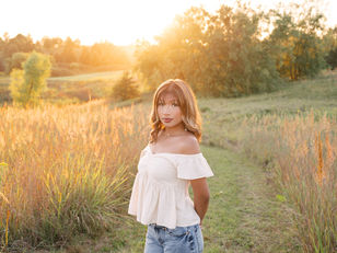 Woman in white off-shoulder top stands in grassy field at sunset, surrounded by tall golden plants, with serene expression.