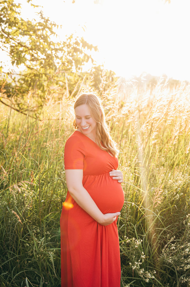 Pregnant woman in a red dress stands in a sunlit field, gently holding her belly and smiling. Tall grass surrounds her in the warm glow.