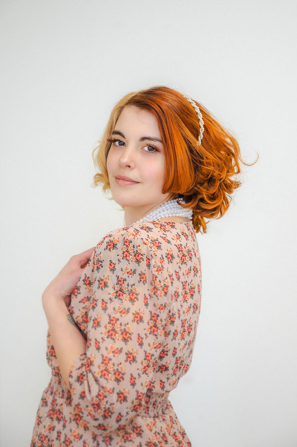 female high school senior with red hair posing in studio with white background wearing pearls and a warm floral dress