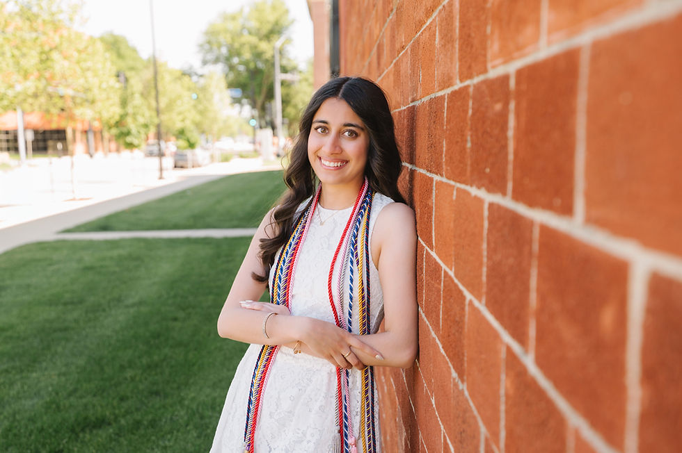 A young woman in a white dress, adorned with graduation cords, leans against a brick wall while smiling confidently on a sunny day.
