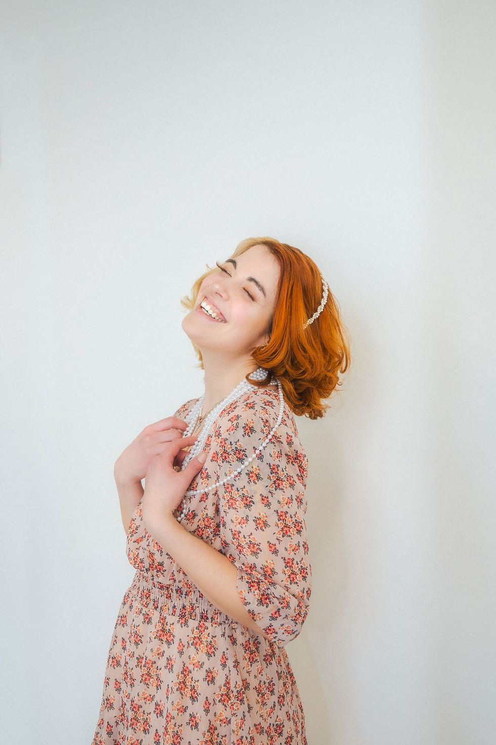female high school senior with red hair posing in studio with white background wearing pearls and a warm floral dress