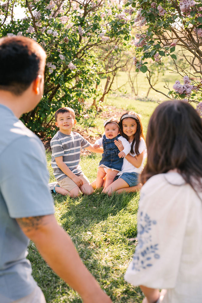 Three children sit on grass surrounded by blooming bushes, with adults in foreground. The mood is joyful, with bright sunlight enhancing the colors.
