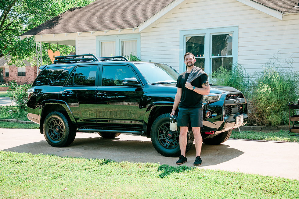 Landscape photo of a shiny black SUV parked in front of a light house with a person standing beside it holding detailing tools