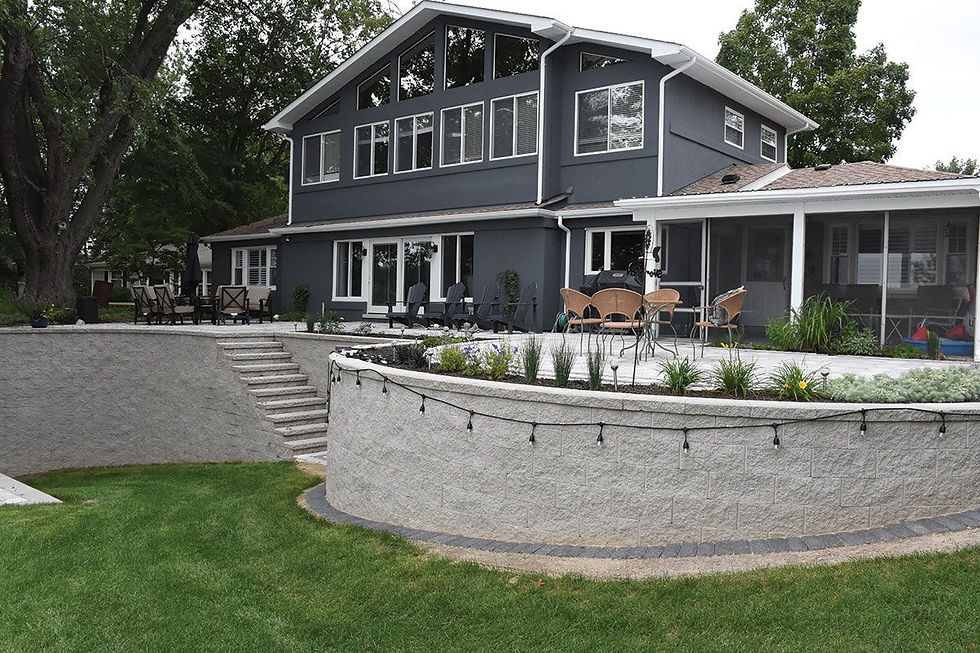 A retaining wall in a backyard, wall is 8"x18" grey with a split face finish. Top of wall is a raised patio and house