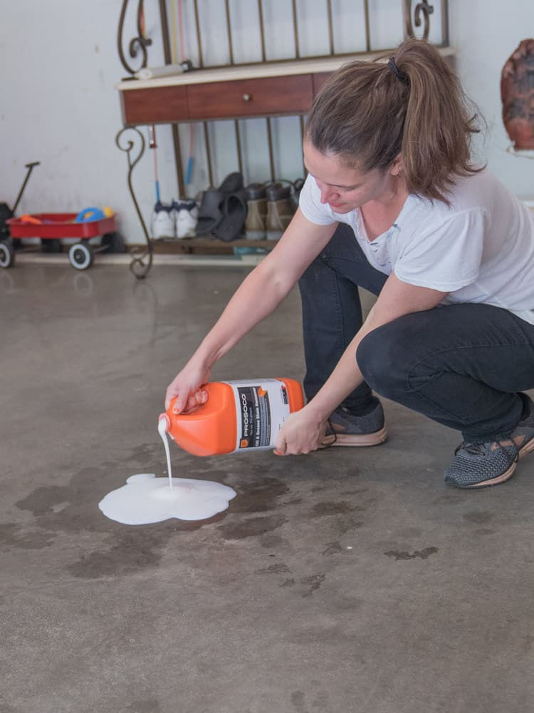 A woman kneeling down in here garage cleaning up an oil spill. She is pouring a white thick substance over the stain.