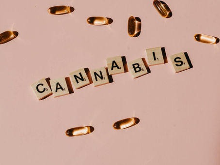 Gold capsules scattered on a pink background with Scrabble tiles spelling "CANNABIS." Soft lighting casts subtle shadows.