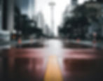 Wet urban street with orange cones, empty under grey skies, flanked by tall buildings and trees. Reflective red road surface adds contrast.