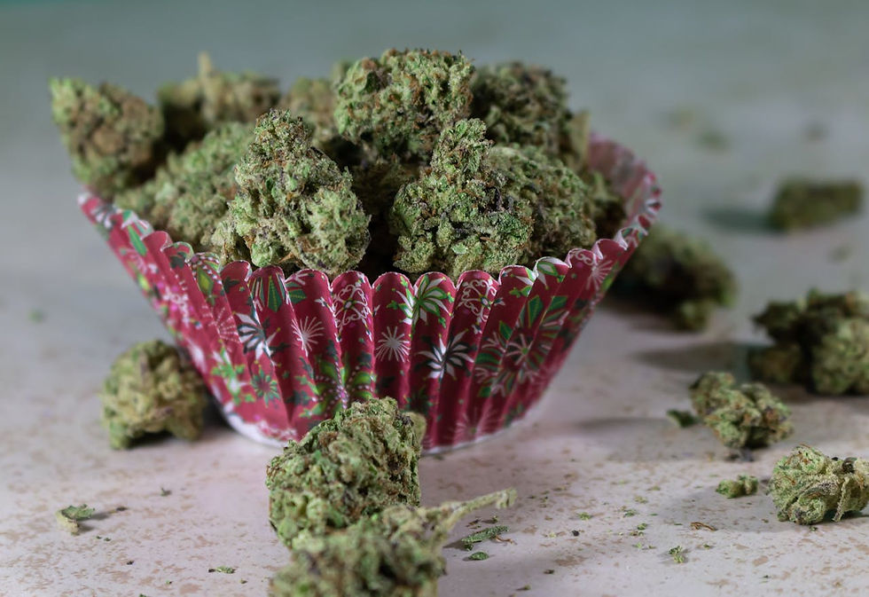 Green cannabis buds in a red, patterned cupcake liner on a light surface. Additional buds scattered around, creating a casual, earthy vibe.