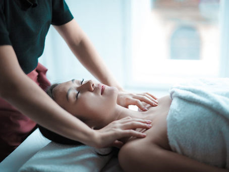 Person receiving a massage in a bright room, covered with a white towel. The masseuse's hands are on their shoulders. Relaxed mood.