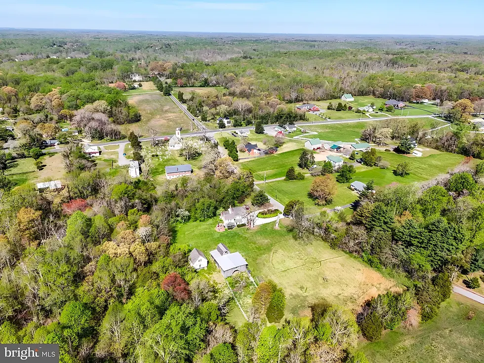 Aerial view of a rural neighborhood with houses, barns, a church with a white steeple, open fields, and dense green trees under a clear blue sky.