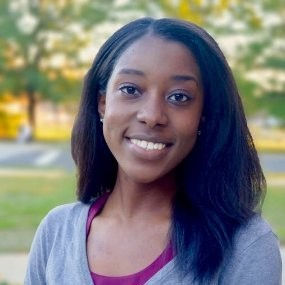 Portrait of a smiling woman with long black hair, standing outside on a sunny day with blurred green trees in the background.