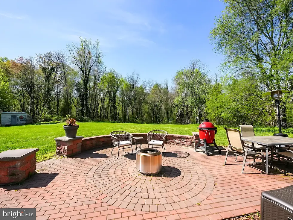 Outdoor patio with circular red brick design, a stainless steel fire pit, wicker chairs with cushions, a red ceramic grill, and a backdrop of grass and tall trees under a clear blue sky.