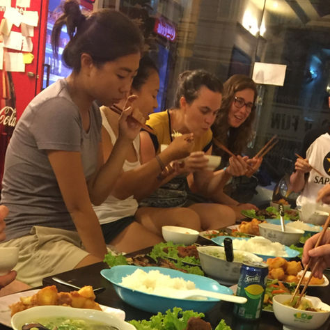 Jonge reiziger zit alleen aan een lange tafel in een hostelkeuken met een kop koffie, omringd door andere reizigers in gesprek