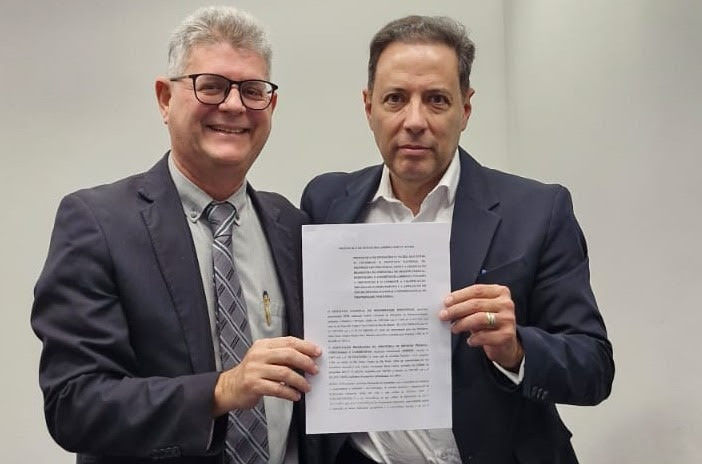 Dois homens de terno, um sorrindo, seguram um documento com texto ilegível diante de parede clara.