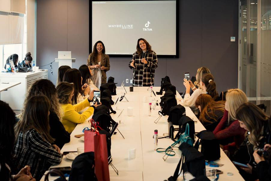 Palestrantes em sala de reunião; tela exibe Maybelline e TikTok. Público sentado à mesa, filmando com celulares.