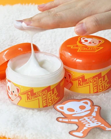 Mão puxando creme branco elástico de potes laranja com desenho de macaco e texto RETINOL CREAM ELASTIC, sobre toalha branca.