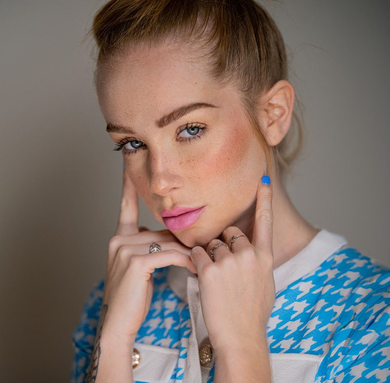 Mulher em close-up com dedos nas bochechas, jaqueta azul xadrez e fundo neutro, expressão calma e intensa.