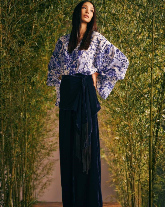Mulher em quimono floral azul e preto posa entre bambus, com expressão serena e olhar ao longe.