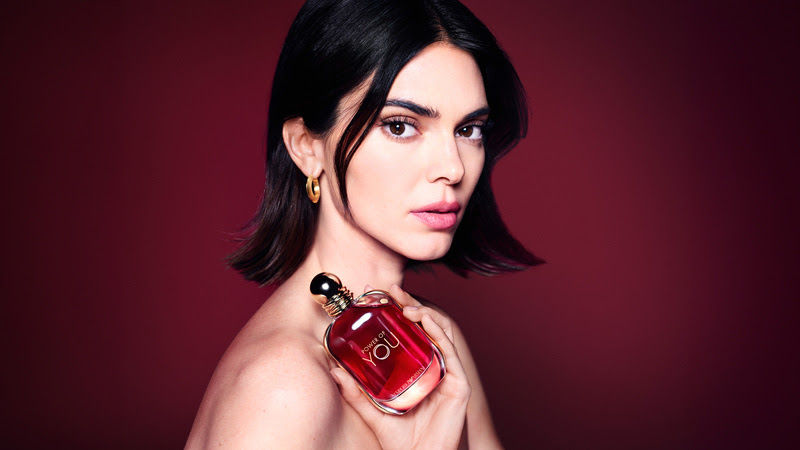 Mulher de cabelo curto segura perfume vermelho com texto you, diante de fundo vinho, expressão séria.