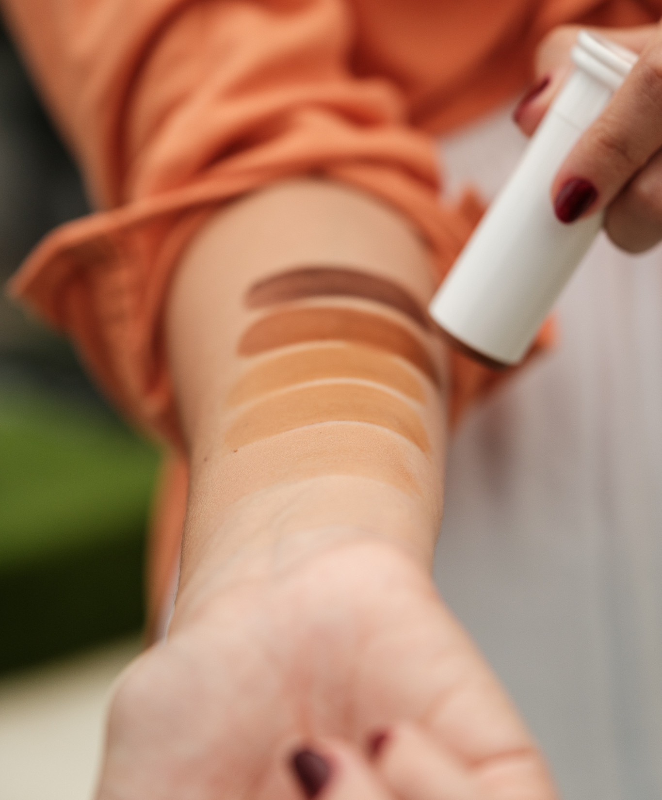 Close-up de braço com amostras de base/bronzer em tons de bege e marrom; mão com esmalte vinho segura um bastão branco.