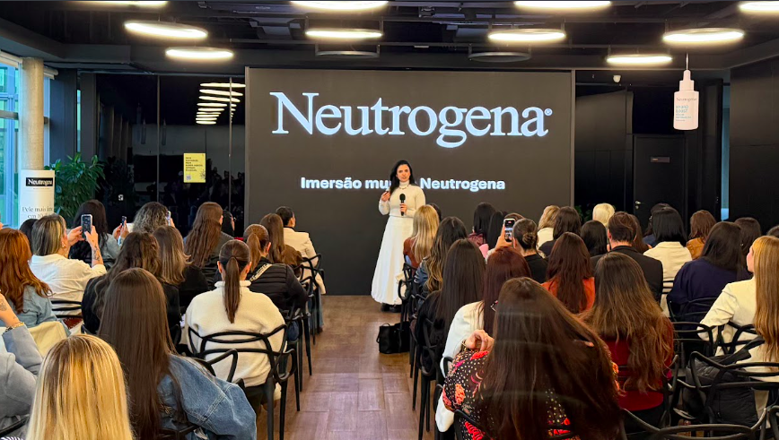 Palestrante de vestido branco fala ao público em evento Neutrogena; tela exibe Neutrogena e Imersão mult Neutrogena.