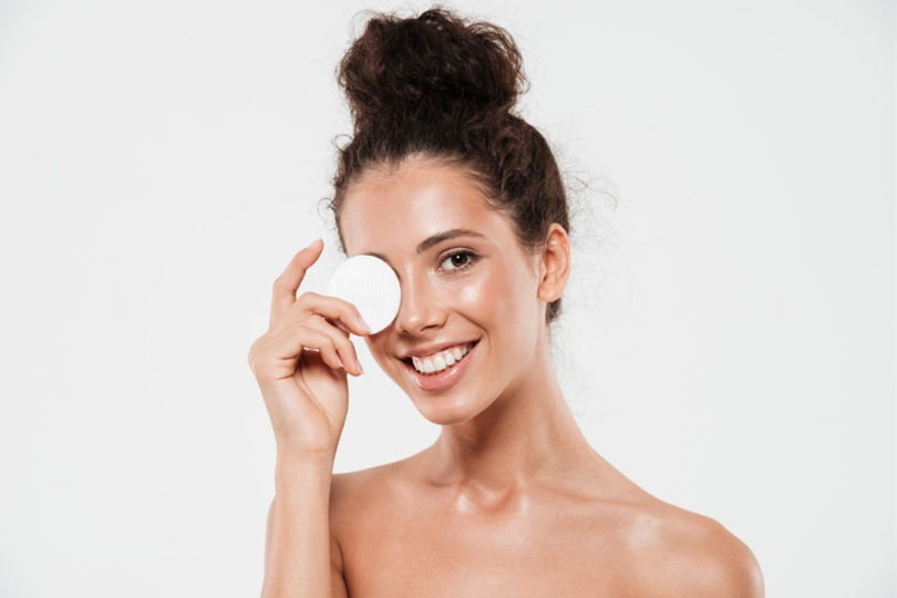 Mulher sorrindo, com cabelo preso, segura um disco de algodão sobre um olho em fundo branco, em clima leve e cuidado facial
