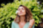 Mulher de olhos fechados com flor branca no cabelo, diante de folhagem verde, em pose serena e relaxada.
