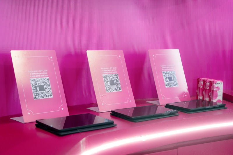 Expositor rosa com três placas de QR code e folhetos, sobre balcão brilhante, em ambiente moderno e delicado.
