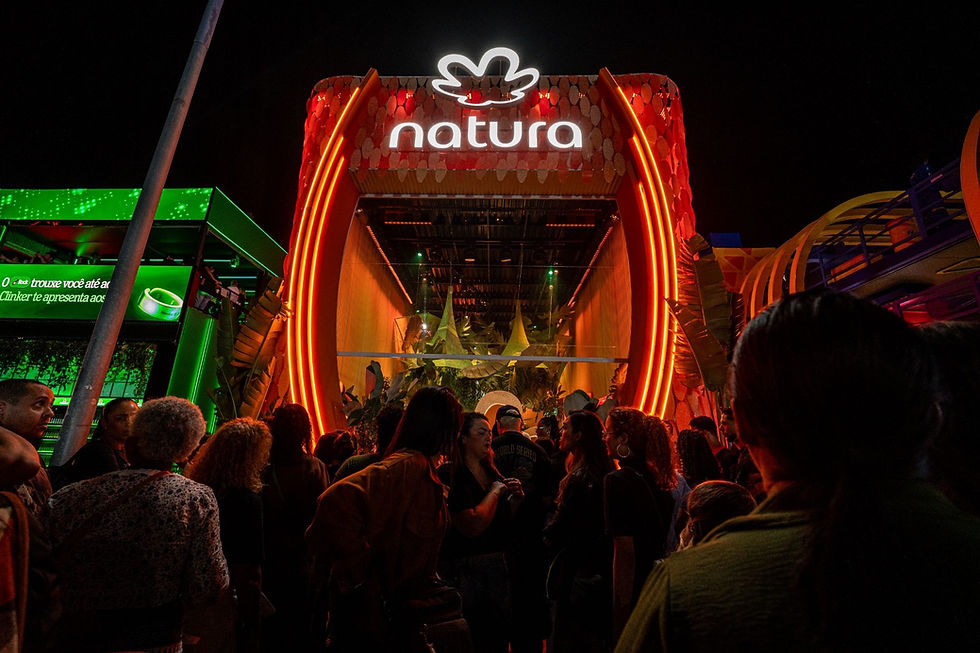 Multidão à noite diante do estande iluminado da Natura, em tons laranja e verde, com ambiente festivo.