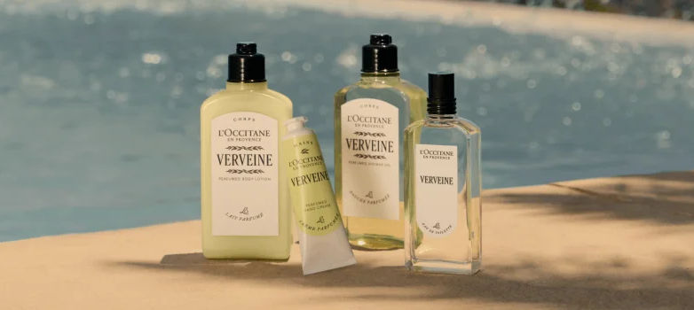 Frascos de produtos L’Occitane Verbena sobre uma borda de piscina, com água azul ao fundo e clima fresco e ensolarado.