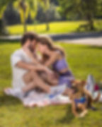 Casal abraçado em piquenique no parque, com cão marrom em primeiro plano; clima romântico e ensolarado.