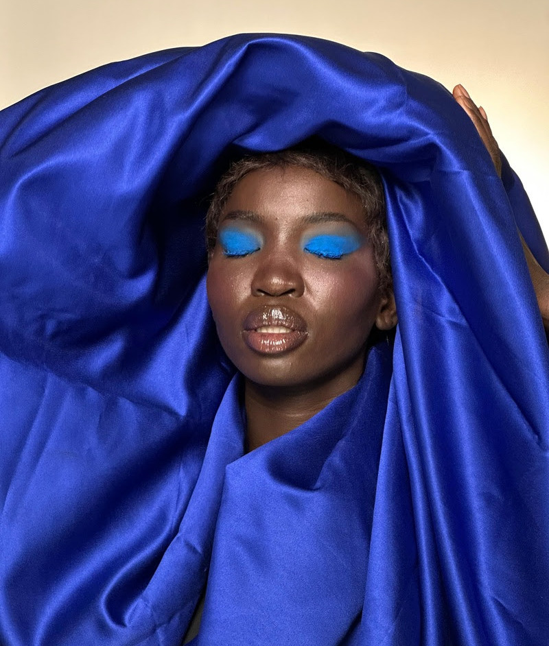 Retrato de mulher com maquiagem azul e tecido azul brilhante cobrindo a cabeça, olhos fechados, fundo bege.