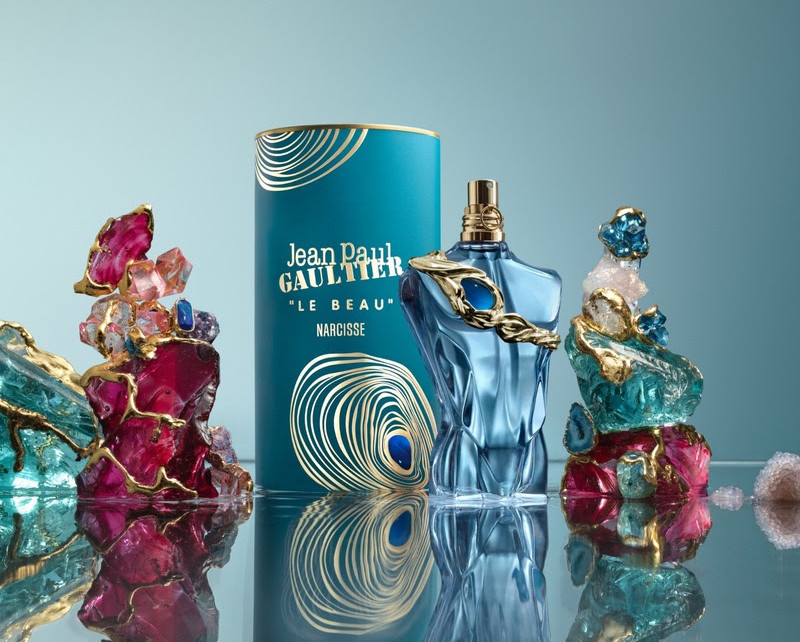 Frasco azul do perfume Jean Paul Gaultier Le Beau Narcisse ao centro, com cristais coloridos sobre fundo azul.