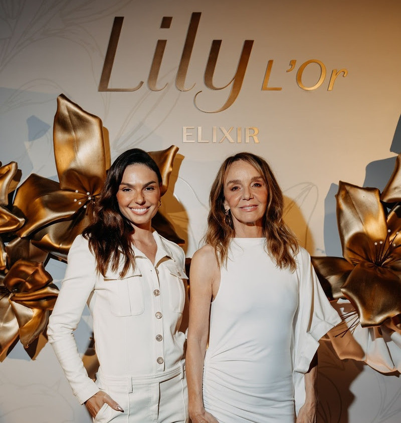 Duas mulheres de branco posam diante do painel Lily L'Or ELIXIR com flores douradas, em evento elegante.