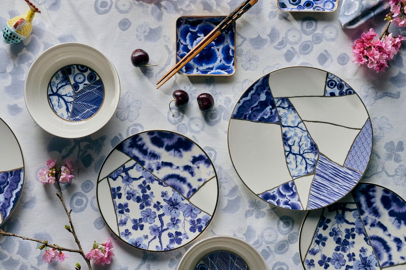 Louças e tigelas azul‑e‑branco sobre mesa floral, com hashi, cerejas e flores rosas, em composição delicada e serena.